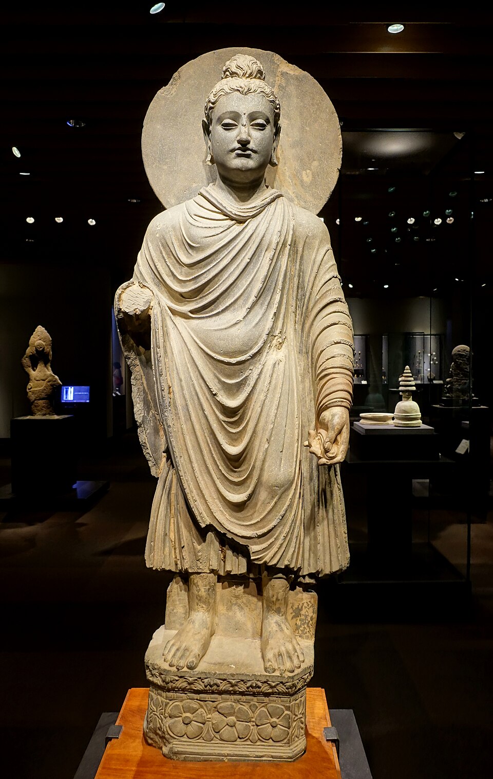 Statue de schiste d'un Bouddha debout, vêtu d'un manteau aux plis profonds, à la manière d'une statue grecque